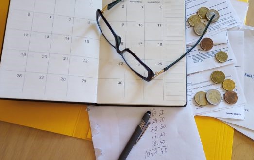 Kalenteri, silmälasit, kolikoita, vihossa laskelma rahojen riittämisestä